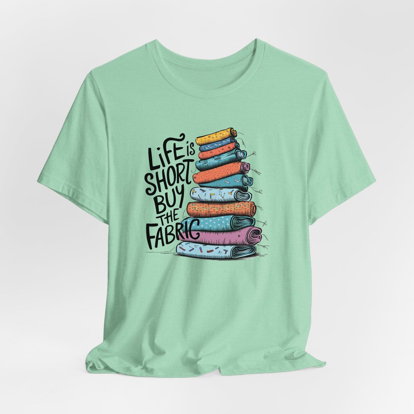 A Heather Mint Short Sleeve T-Shirt with a illustration of colorful fabric rolls with bold "Life is Short Buy the Fabric" text