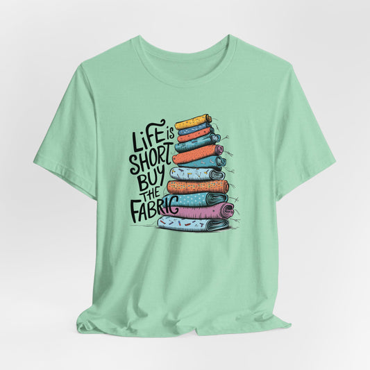 A Heather Mint Short Sleeve T-Shirt with a illustration of colorful fabric rolls with bold "Life is Short Buy the Fabric" text