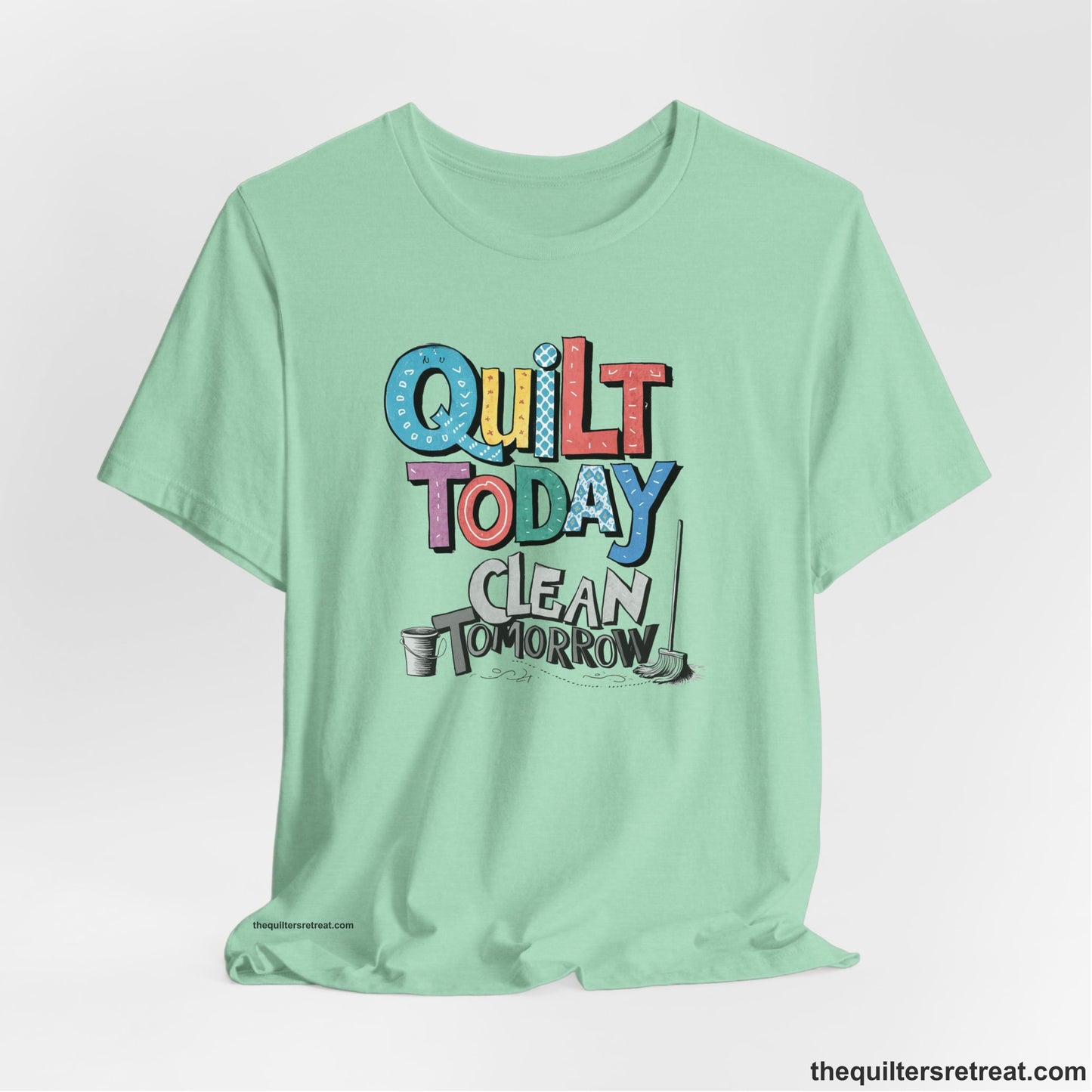 A Heather Mint Short Sleeve T-Shirt with a playful "Quilt Today Clean Tomorrow" text with colorful patterns and cleaning tools illustration