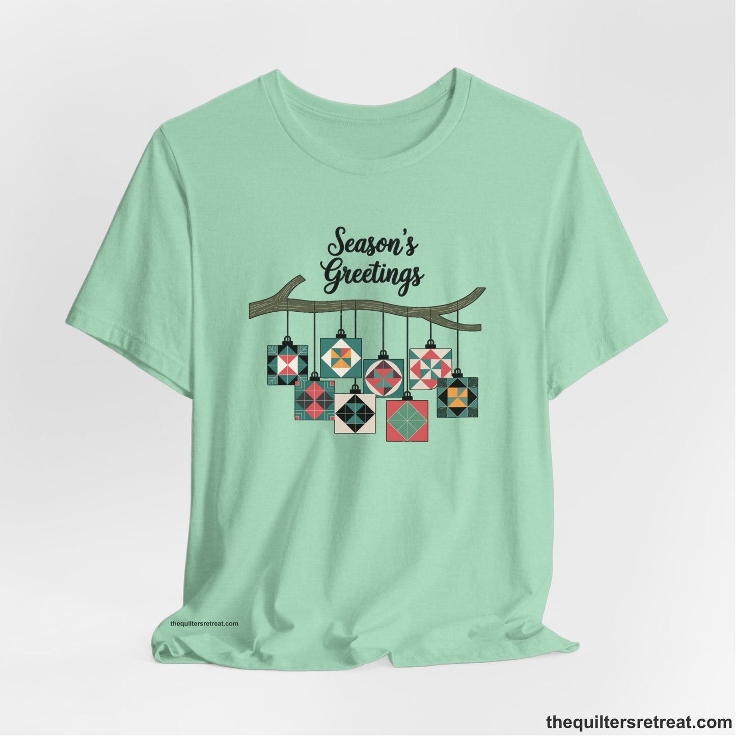 A Heather Mint Short Sleeve T-Shirt with a season's greetings text above colorful quilt block ornaments on tree branch in rustic style