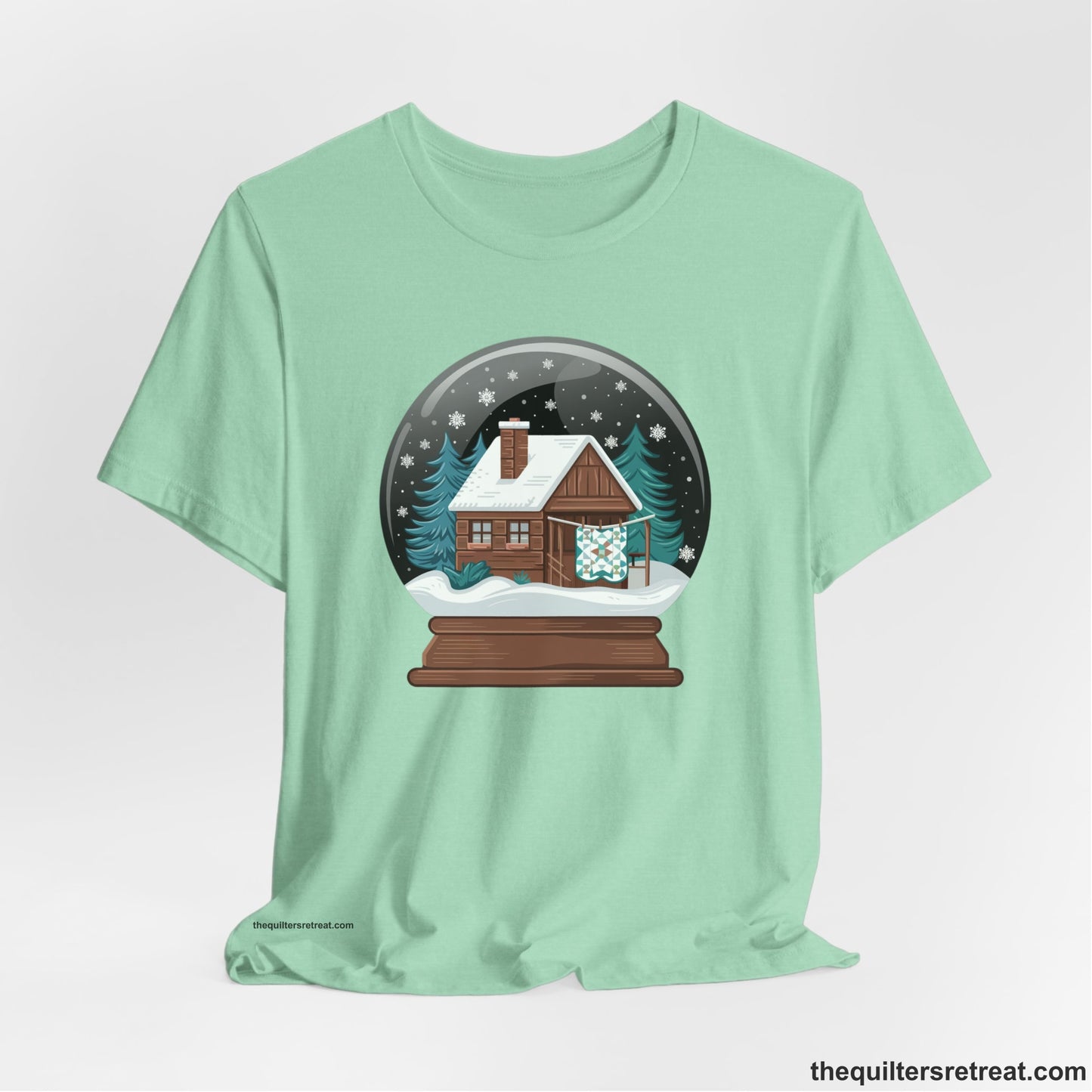 A Heather Mint Short Sleeve T-Shirt with a cabin in a snow globe with snowflakes and trees, in a cozy rustic style