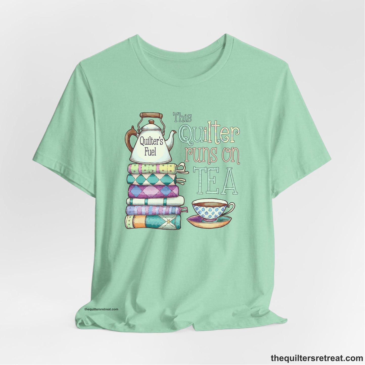 A Heather Mint Short Sleeve T-Shirt with a stack of colorful quilts with teapot and cup, whimsical "This Quilter Runs on Tea" text