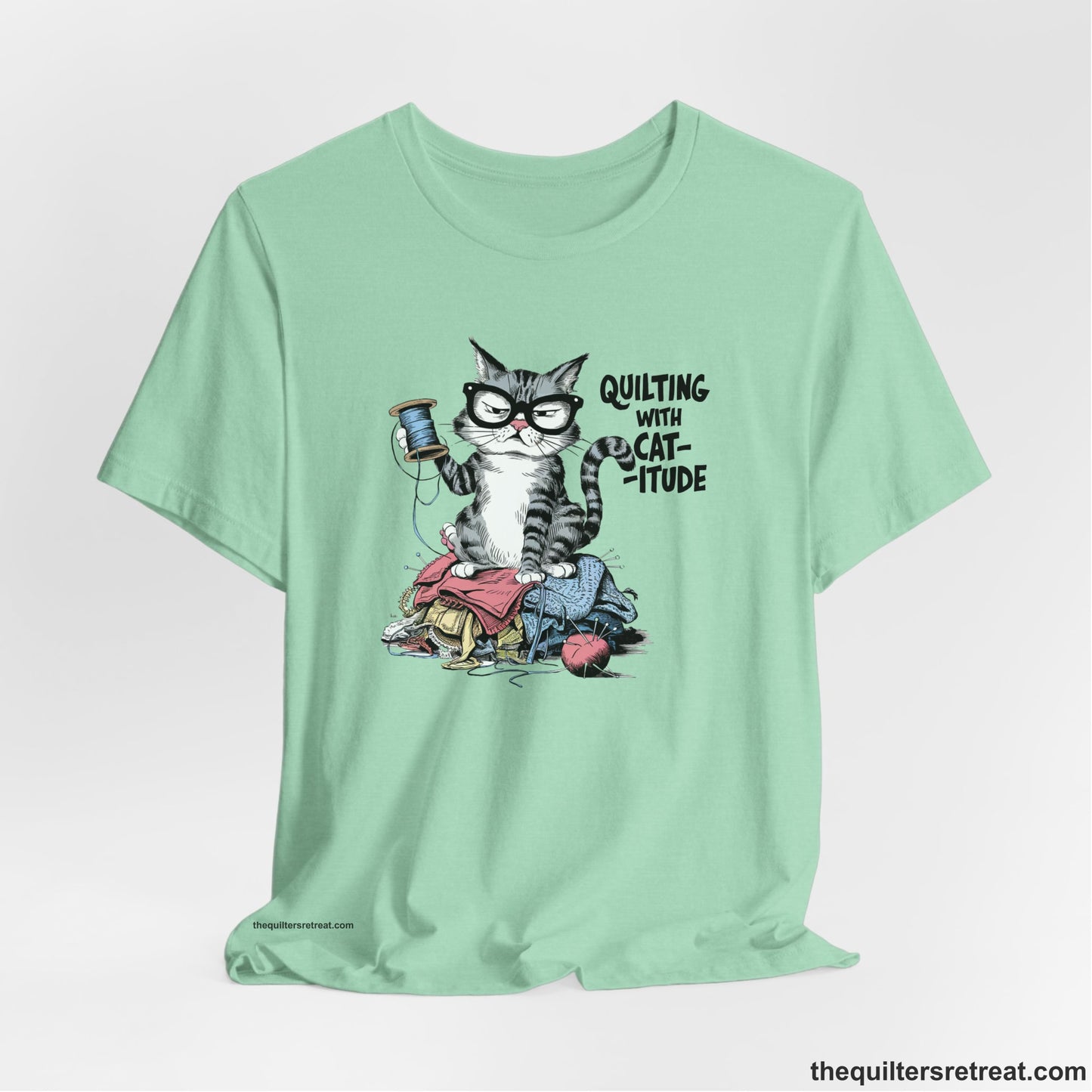 A Heather Mint Short Sleeve T-Shirt with a grumpy cat holding thread spool atop fabric pile with "Quilting with Cat-itude" text