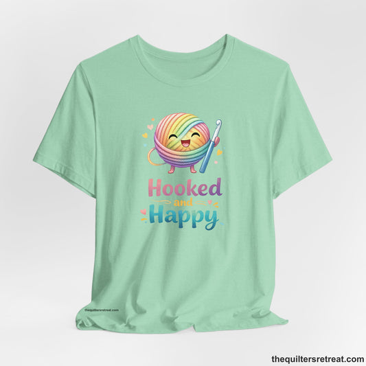 Crochet Gift: A Heather Mint Short Sleeve T-Shirt with Smiling rainbow yarn ball with crochet hook, hearts, and "Hooked and Happy" text, whimsical style.