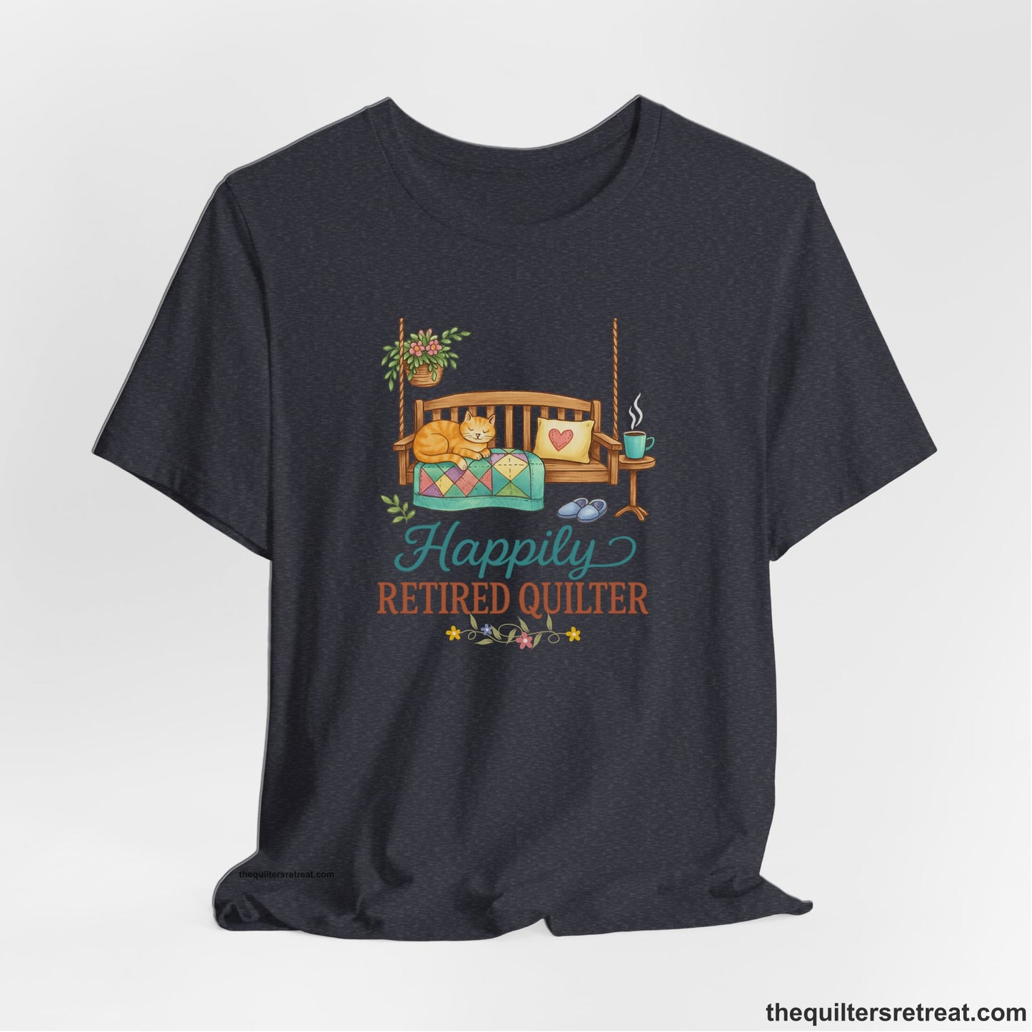 A Heather Navy Short Sleeve T-Shirt with a hanging bench with cozy cat and quilt, surrounded by plants, "Happily Retired Quilter" text