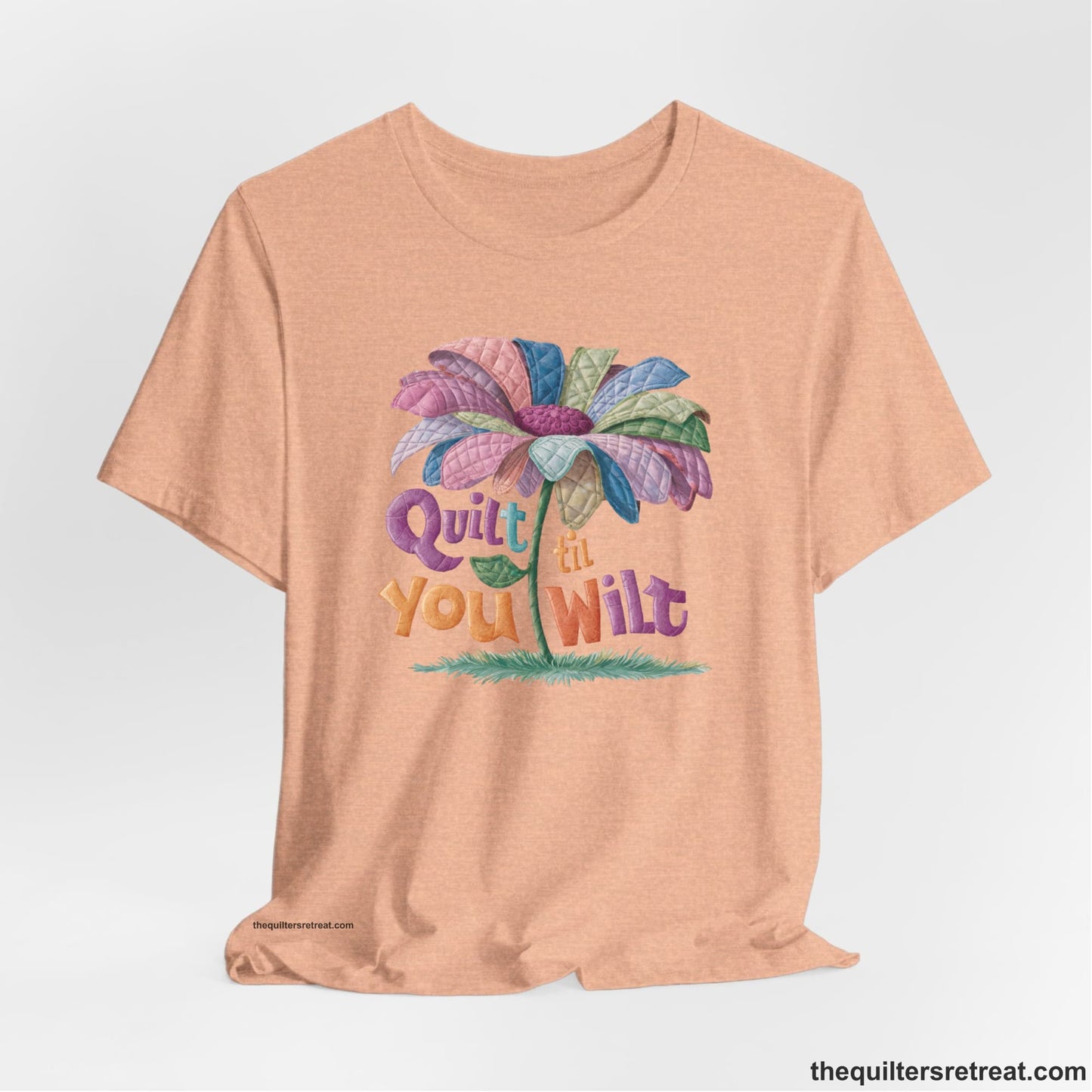 A Heather Peach Short Sleeve T-Shirt with a whimsical quilted flower with bold "Quilt til You Wilt" text
