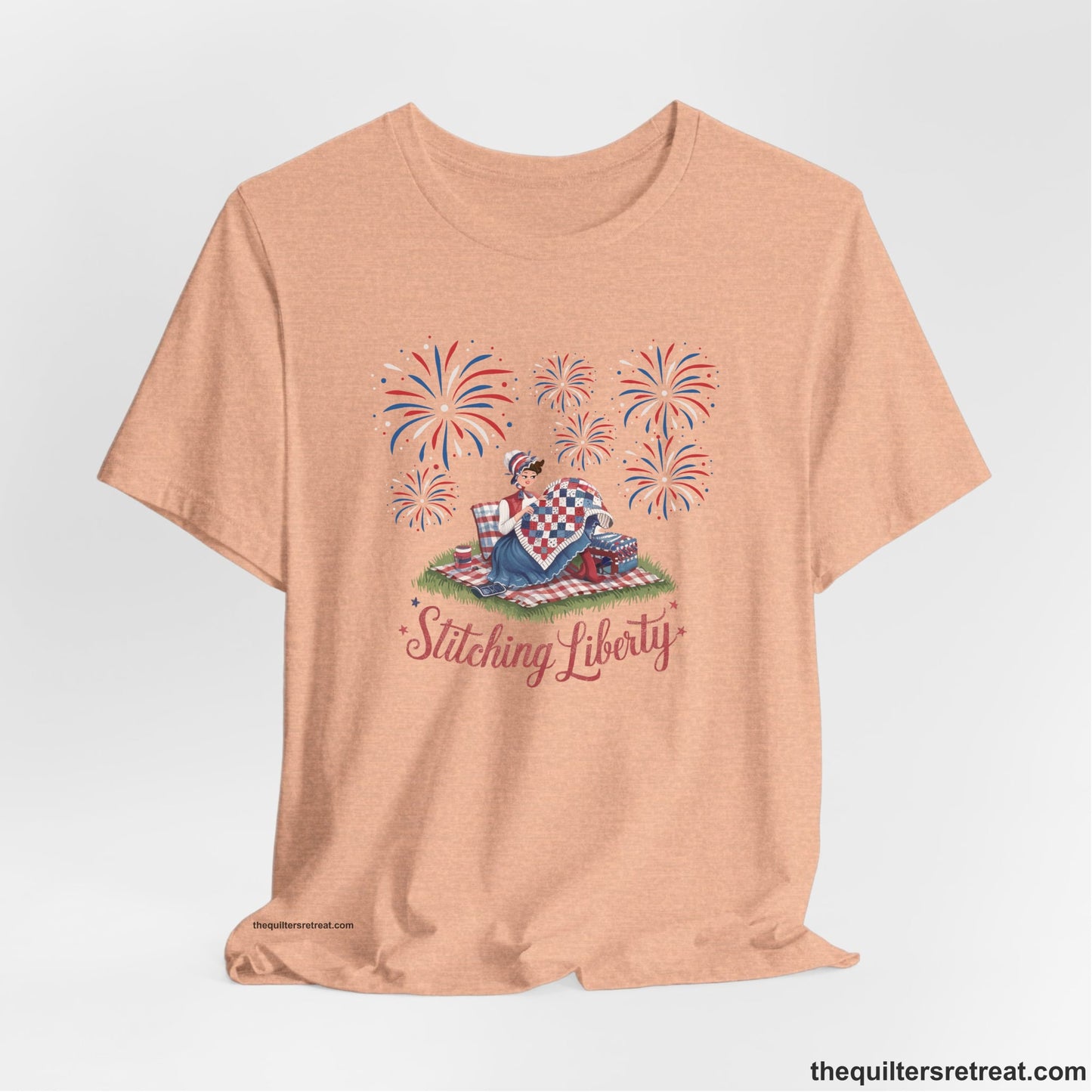 A Heather Peach Short Sleeve T-Shirt with a vintage illustration of woman quilting with fireworks and "Stitching Liberty" text in patriotic style