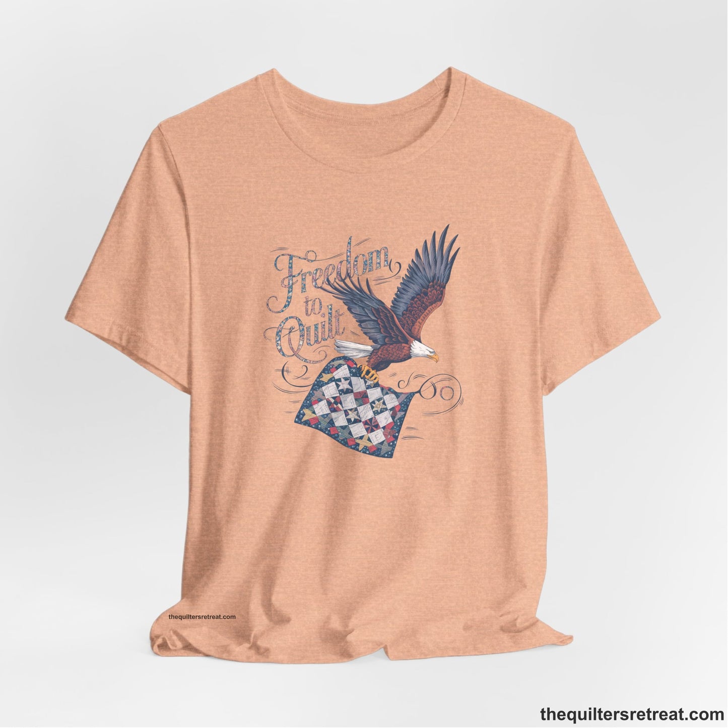 A Heather Peach Short Sleeve T-Shirt with a freedom to quilt text with an eagle carrying a colorful patchwork quilt in vintage style