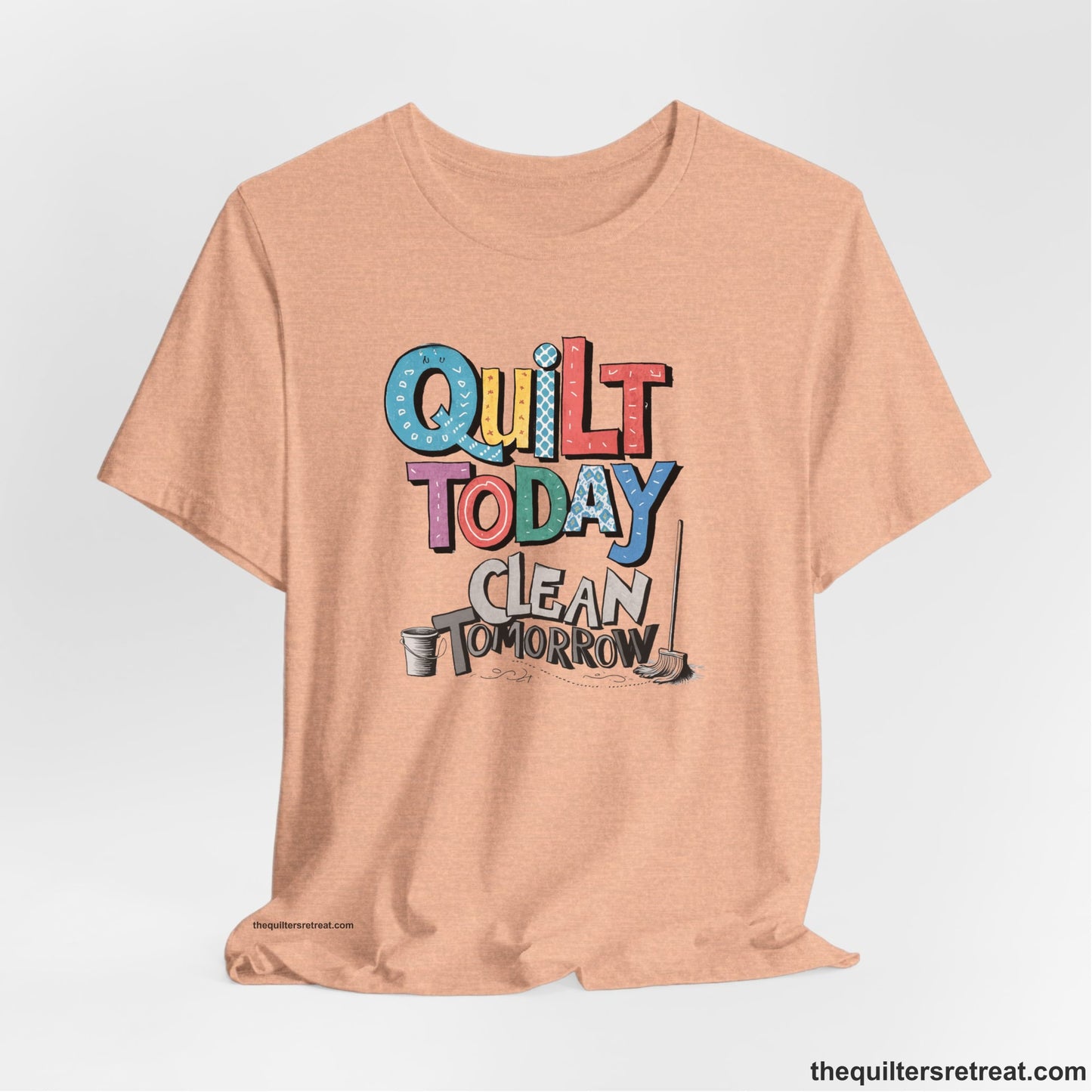 A Heather Peach Short Sleeve T-Shirt with a playful "Quilt Today Clean Tomorrow" text with colorful patterns and cleaning tools illustration