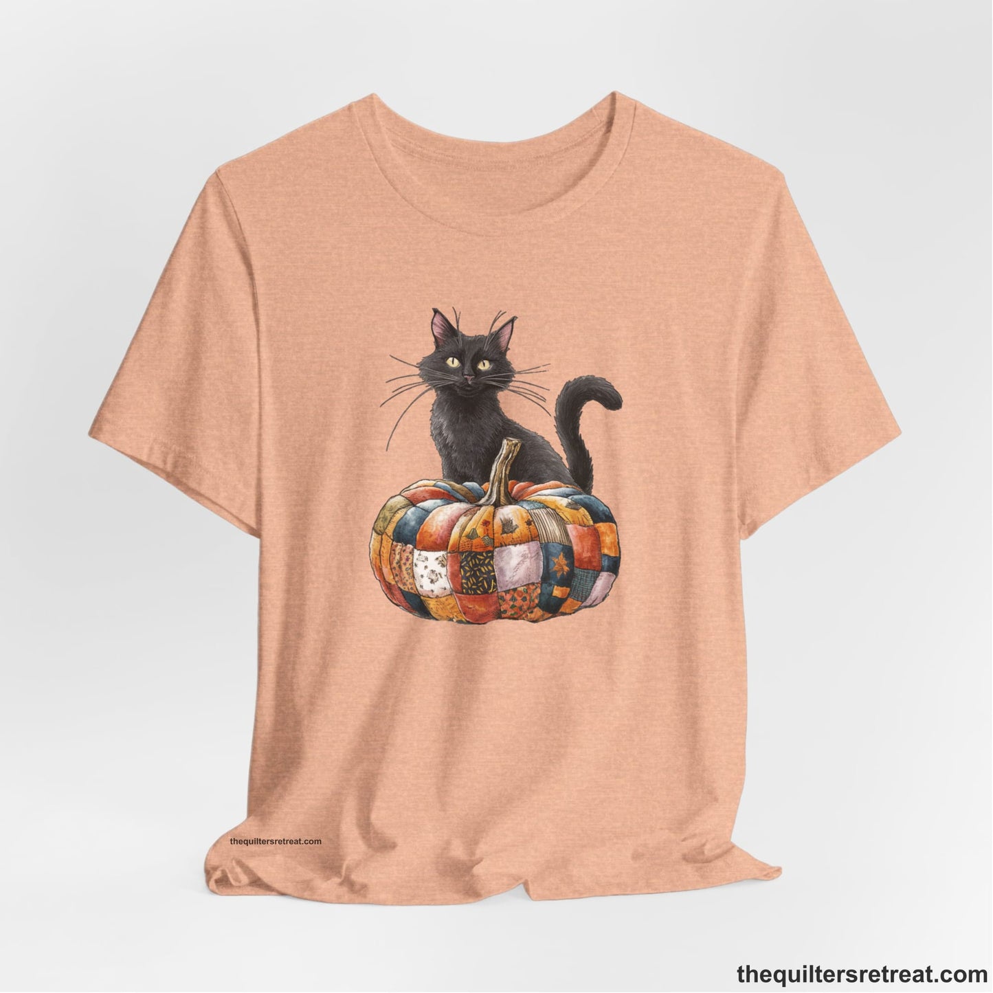 A Heather Peach Short Sleeve T-Shirt with a black cat with a quilted patchwork pumpkin in a whimsical autumn style