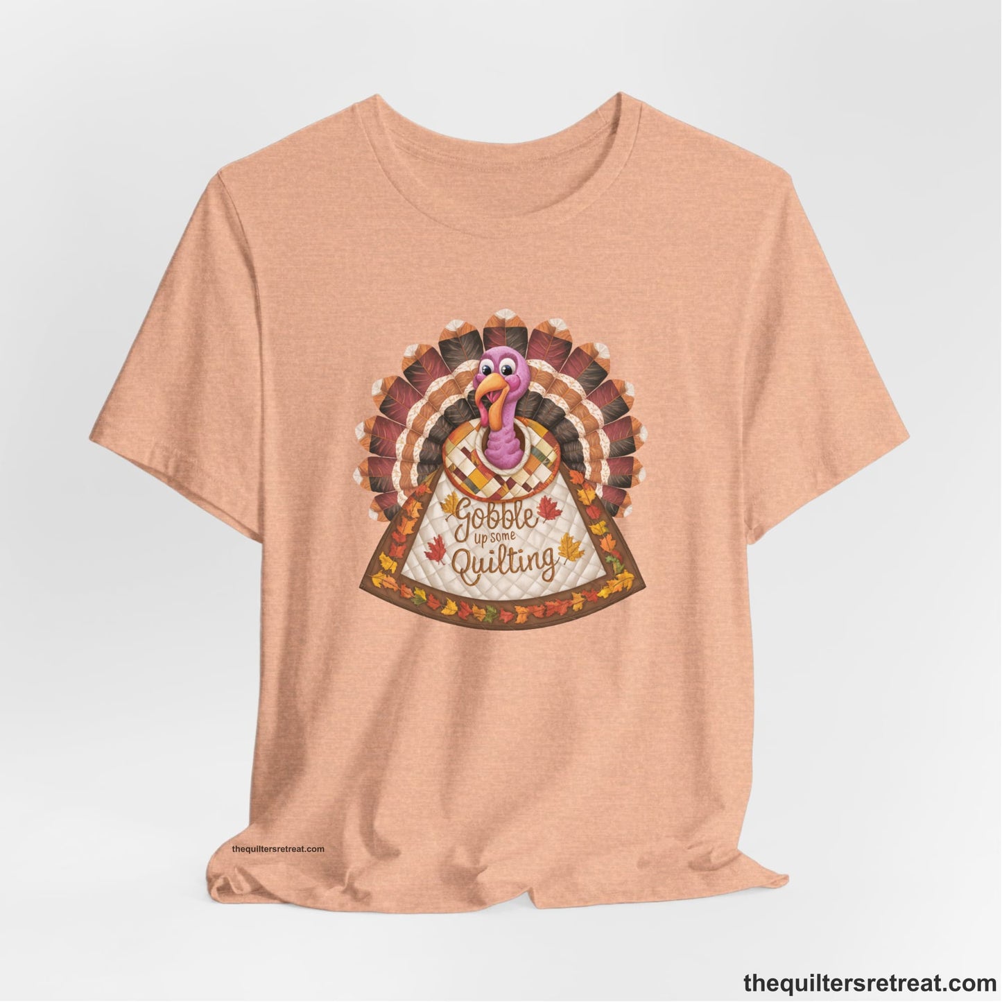 A Heather Peach Short Sleeve T-Shirt with a colorful turkey with quilted feathers and "Gobble Up Some Quilting" text in a playful style