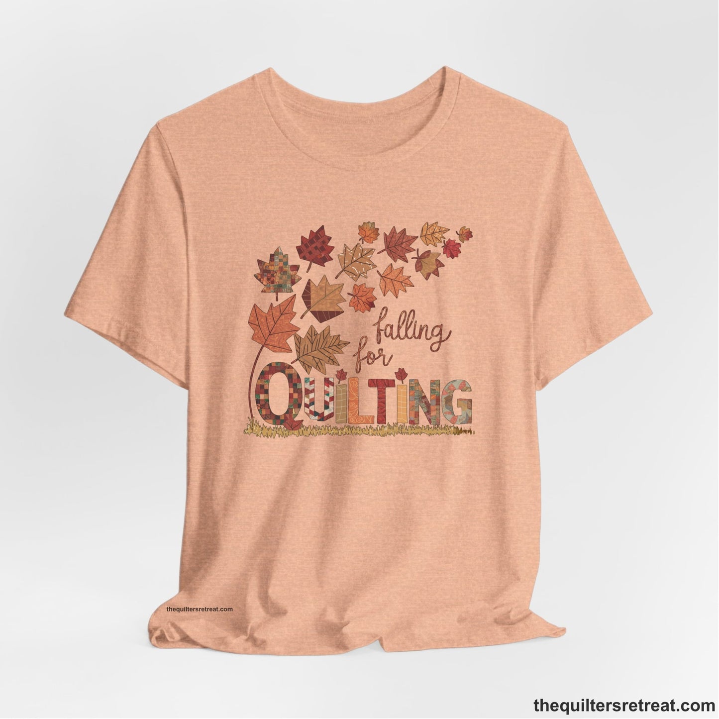 A Heather Peach Short Sleeve T-Shirt with a falling autumn leaves and "Falling for Quilting" text in a patchwork style