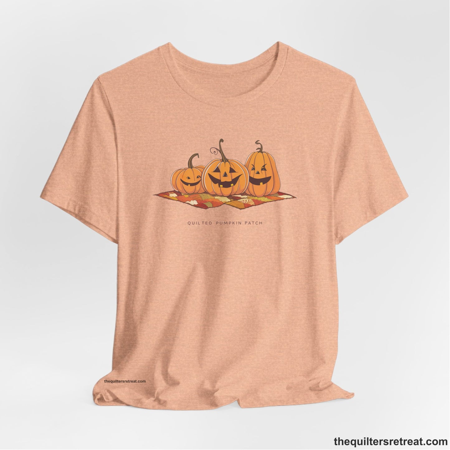 A Heather Peach Short Sleeve T-Shirt with a playful jack-o'-lantern trio on a quilt with "Quilted Pumpkin Patch" text