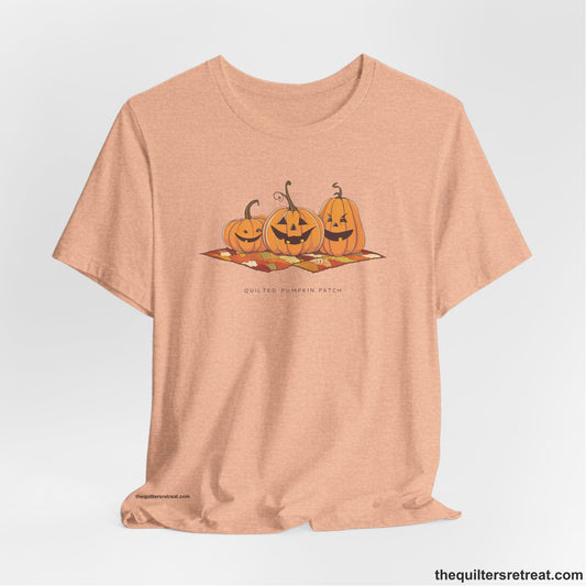 A Heather Peach Short Sleeve T-Shirt with a playful jack-o'-lantern trio on a quilt with "Quilted Pumpkin Patch" text