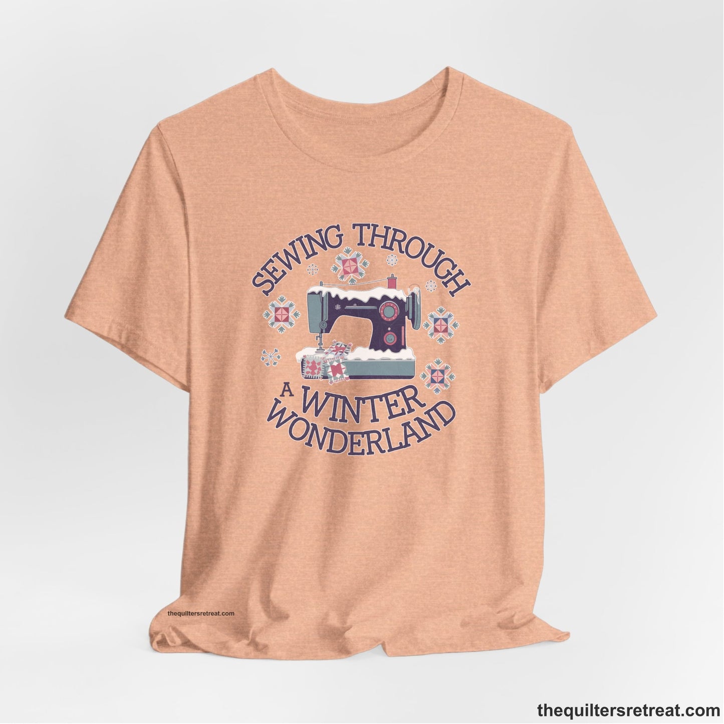 A Heather Peach Short Sleeve T-Shirt with a sewing machine covered in snow with festive quilting elements and "Sewing Through a Winter Wonderland" text