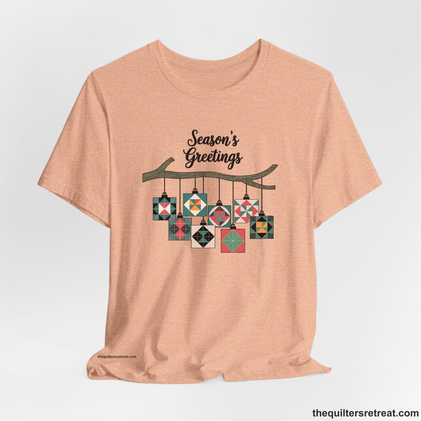 A Heather Peach Short Sleeve T-Shirt with a season's greetings text above colorful quilt block ornaments on tree branch in rustic style