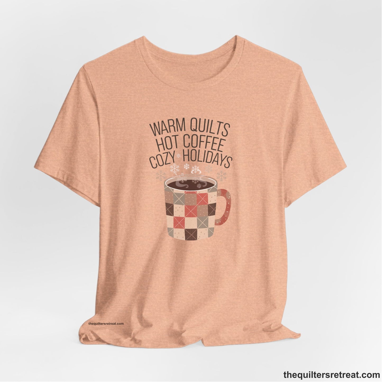 A Heather Peach Short Sleeve T-Shirt with a warm quilts hot coffee cozy holidays text with a patchwork mug and snowflakes