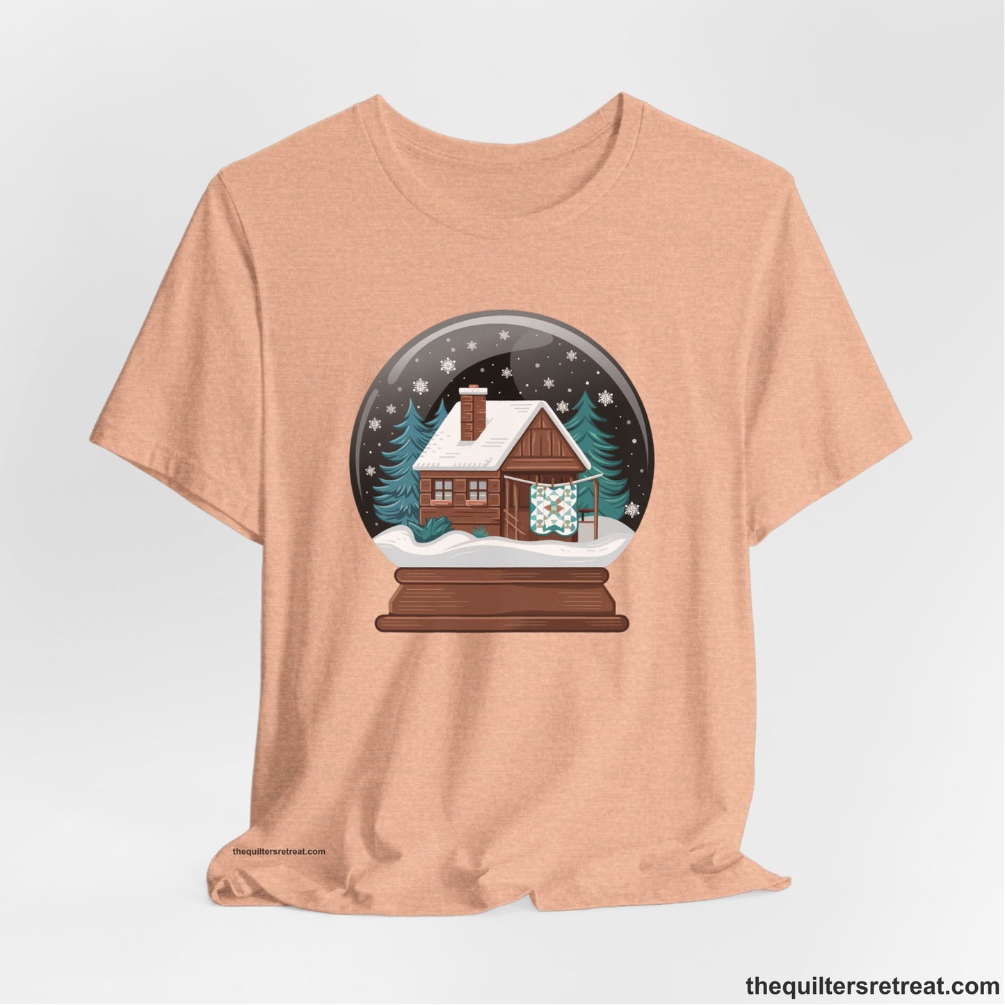 A Heather Peach Short Sleeve T-Shirt with a cabin in a snow globe with snowflakes and trees, in a cozy rustic style