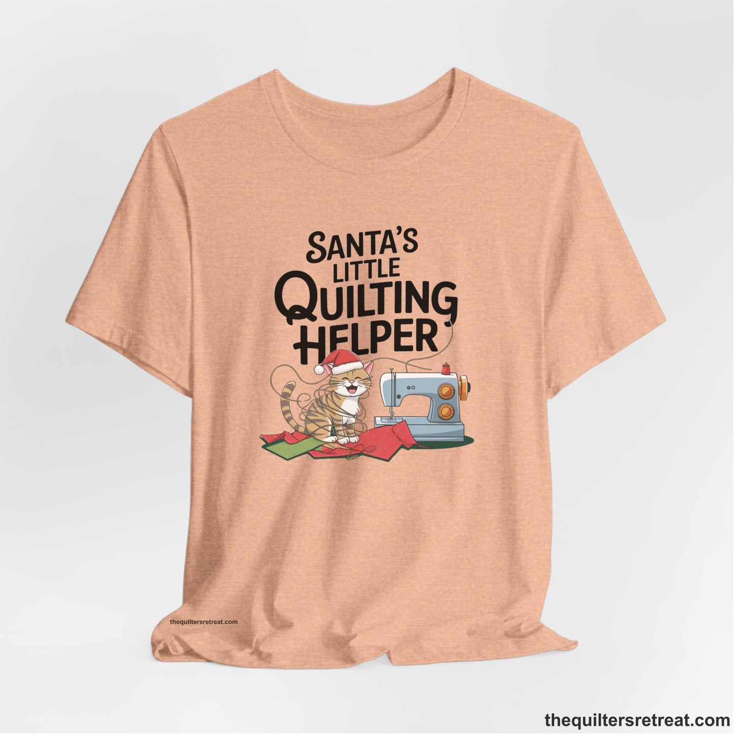 A Heather Peach Short Sleeve T-Shirt with a santa-hat-wearing kitten with a sewing machine and "Santa's Little Quilting Helper" text in playful style