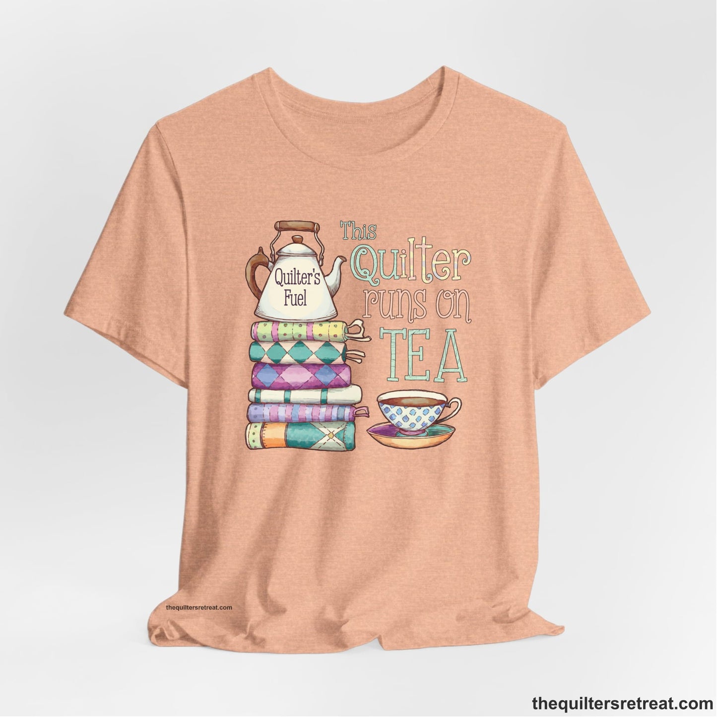 A Heather Peach Short Sleeve T-Shirt with a stack of colorful quilts with teapot and cup, whimsical "This Quilter Runs on Tea" text