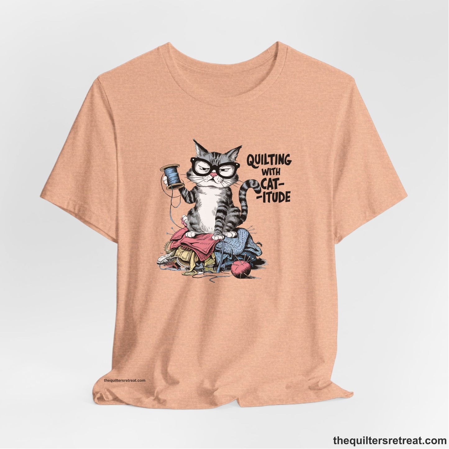 A Heather Peach Short Sleeve T-Shirt with a grumpy cat holding thread spool atop fabric pile with "Quilting with Cat-itude" text