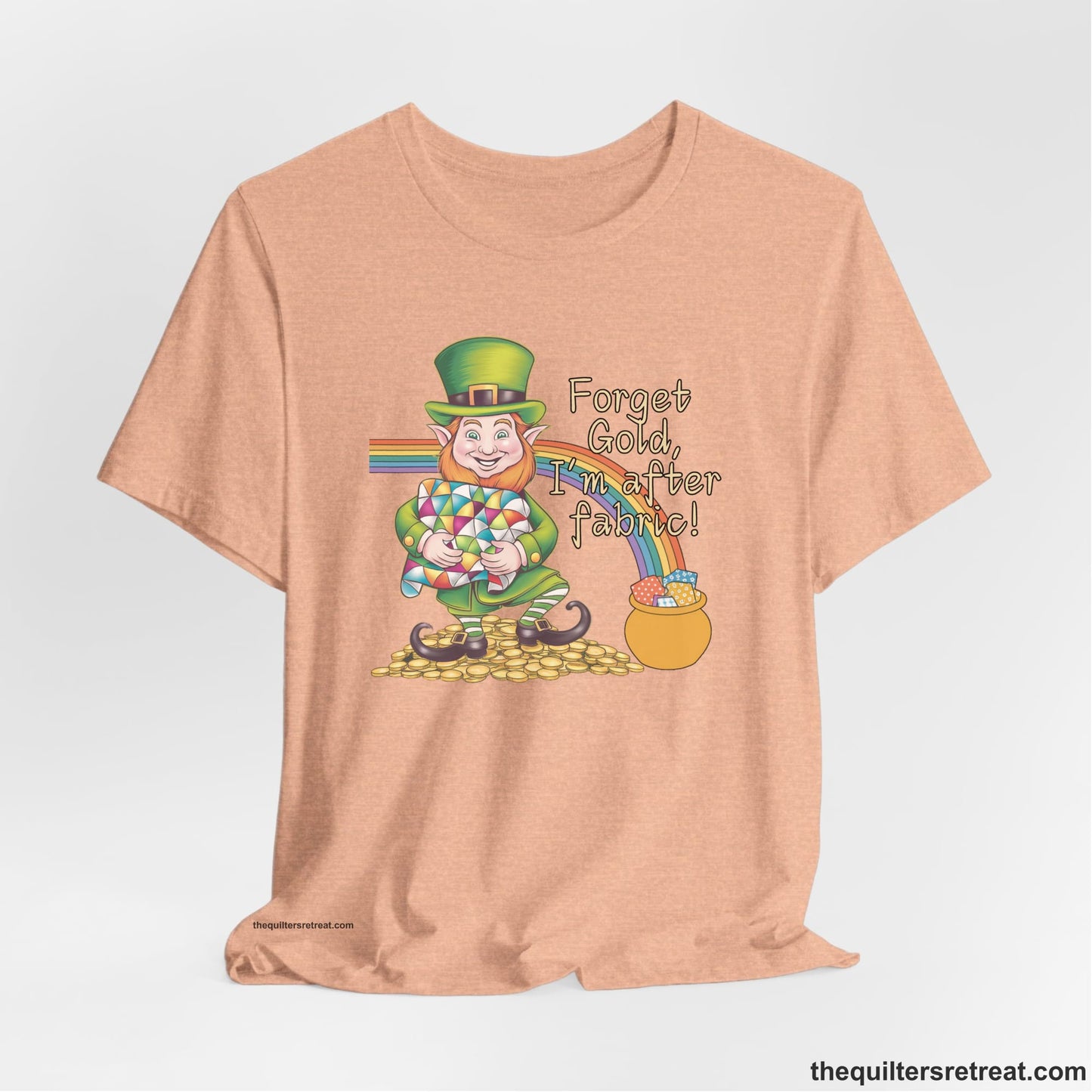 A Heather Peach Short Sleeve T-Shirt with a leprechaun with colorful quilt and rainbow, whimsical "Forget Gold, I'm after fabric!" text