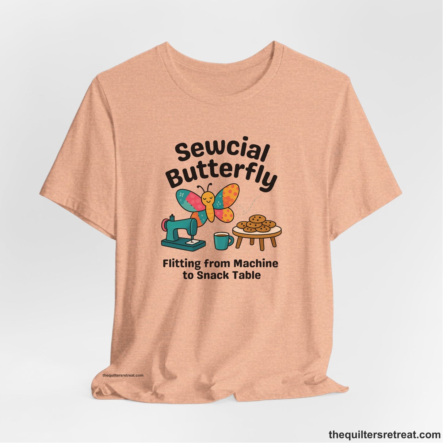 A Heather Peach Short Sleeve T-Shirt with a playful butterfly near sewing machine with cookies and coffee doodle design