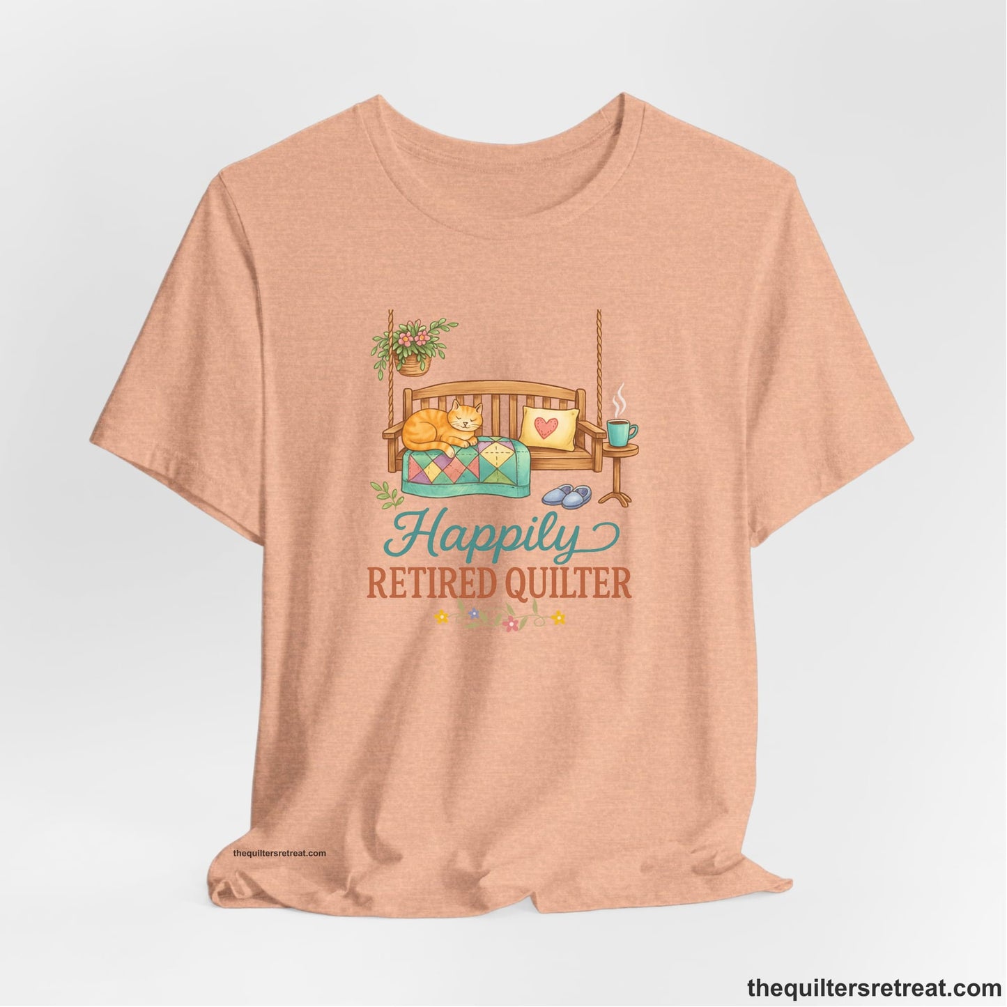 A Heather Peach Short Sleeve T-Shirt with a hanging bench with cozy cat and quilt, surrounded by plants, "Happily Retired Quilter" text
