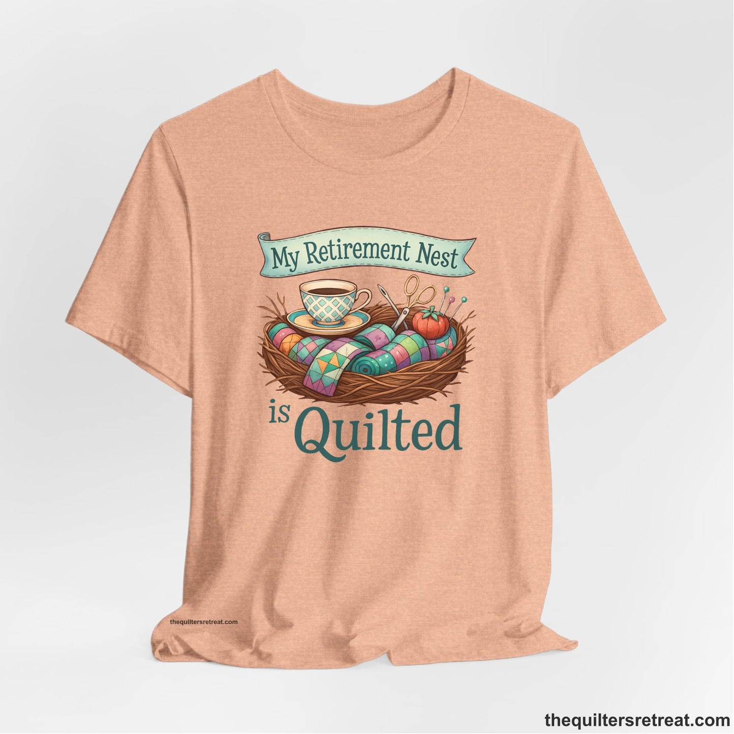 A Heather Peach Short Sleeve T-Shirt with a My Retirement Nest is Quilted text with a cozy sewing elements illustration