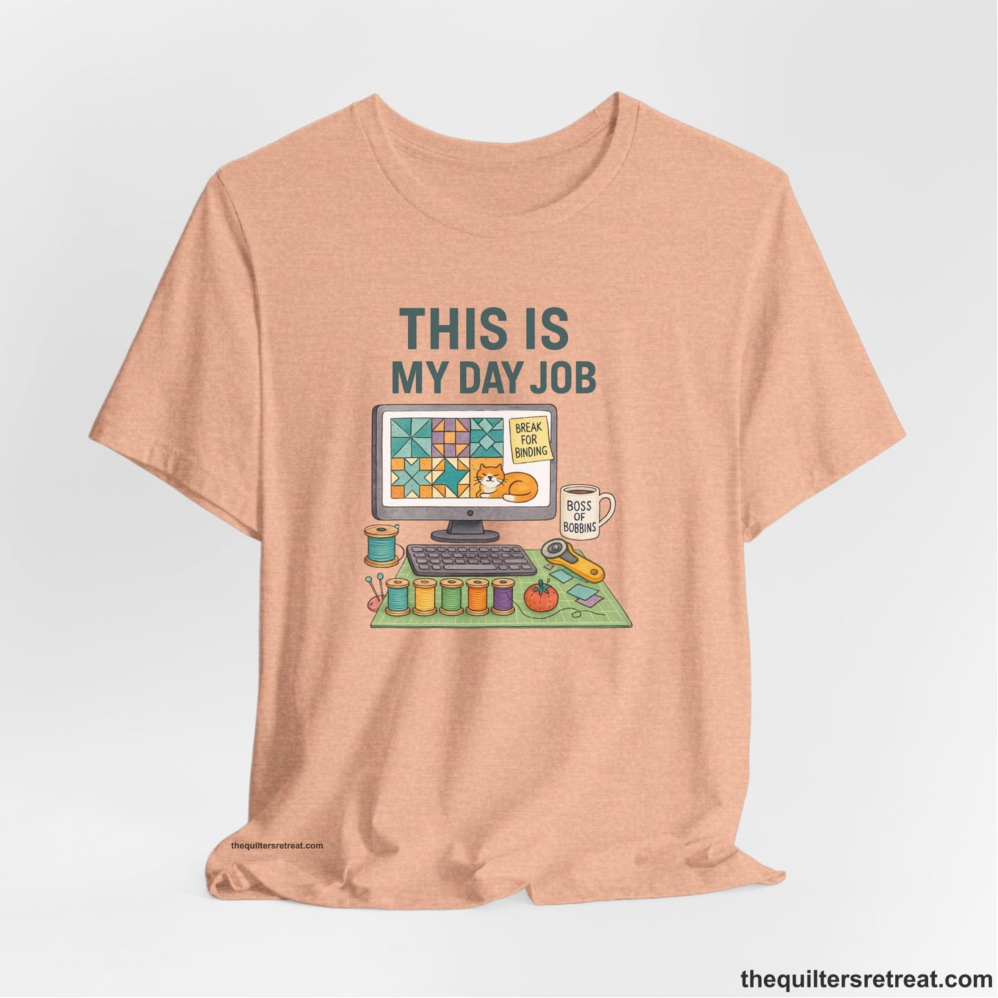A Heather Peach Short Sleeve T-Shirt with a computer quilt design, cat illustration, "THIS IS MY DAY JOB" text with sewing tools, playful style