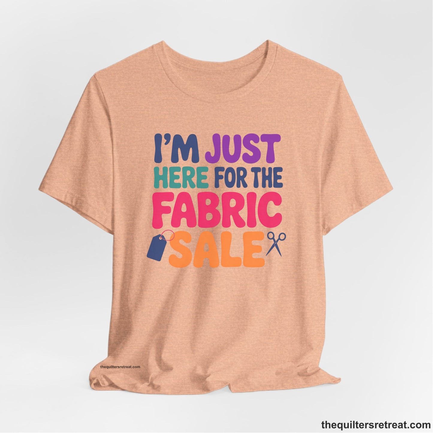 A Heather Peach Short Sleeve T-Shirt with a playful "I'm Just Here for the Fabric Sale" text with colorful letters and sewing icons