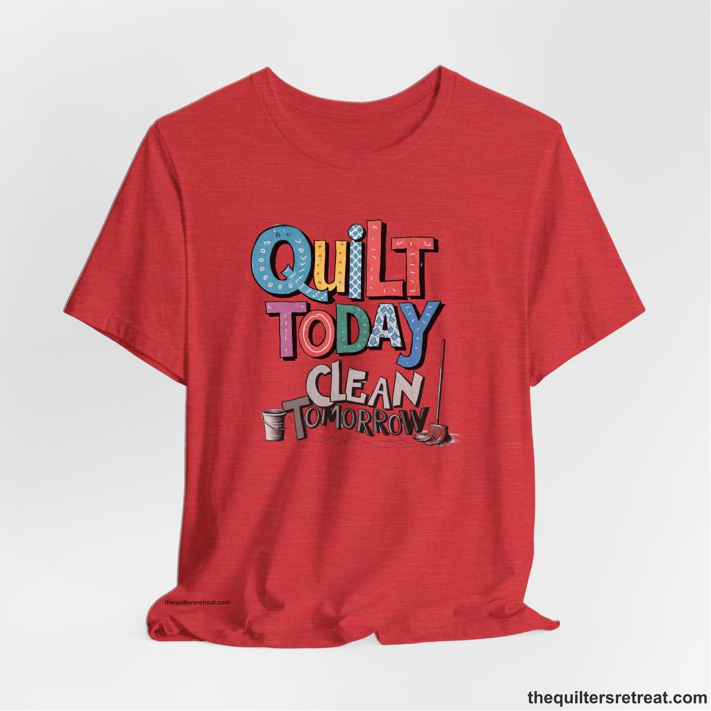 A Heather Red Short Sleeve T-Shirt with a playful "Quilt Today Clean Tomorrow" text with colorful patterns and cleaning tools illustration