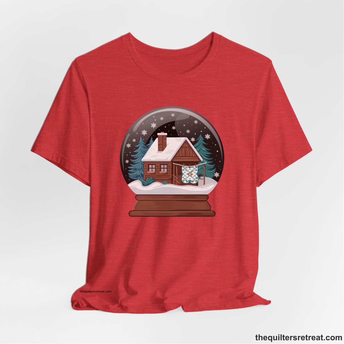 A Heather Red Short Sleeve T-Shirt with a cabin in a snow globe with snowflakes and trees, in a cozy rustic style