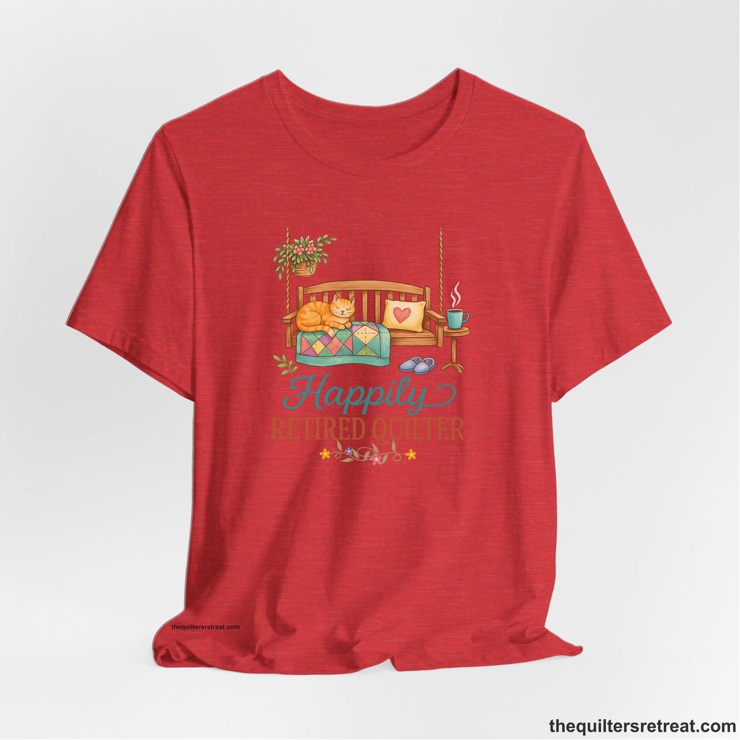 A Heather Red Short Sleeve T-Shirt with a hanging bench with cozy cat and quilt, surrounded by plants, "Happily Retired Quilter" text