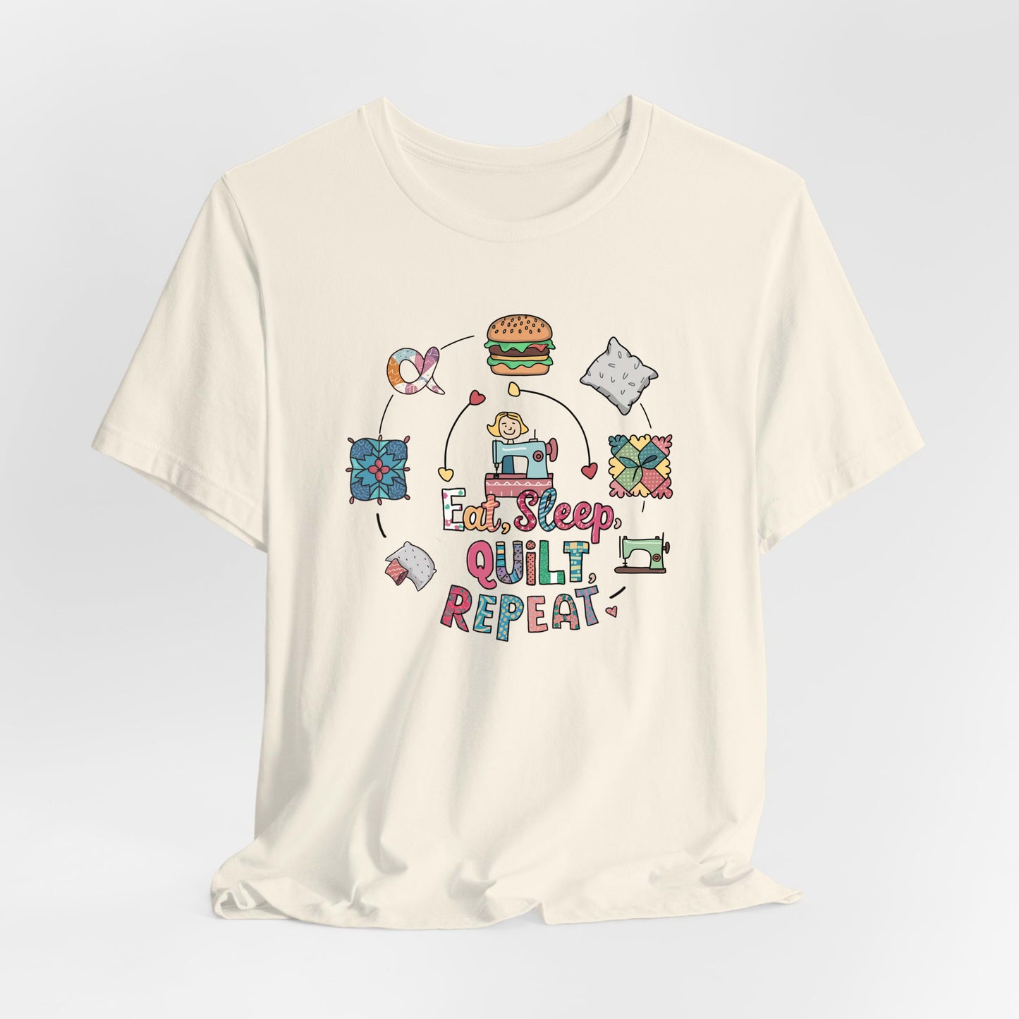 A Natural Short Sleeve T-Shirt with a cute quilting elements with "Eat, Sleep, Quilt, Repeat" text in playful style