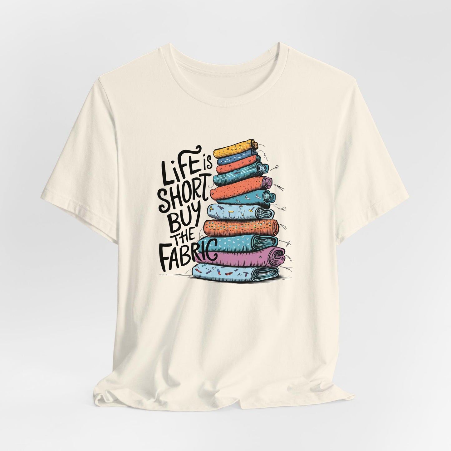 A Natural Short Sleeve T-Shirt with a illustration of colorful fabric rolls with bold "Life is Short Buy the Fabric" text