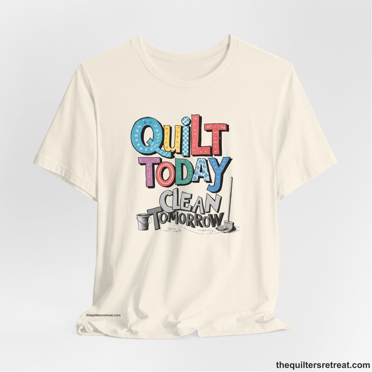A Natural Short Sleeve T-Shirt with a playful "Quilt Today Clean Tomorrow" text with colorful patterns and cleaning tools illustration