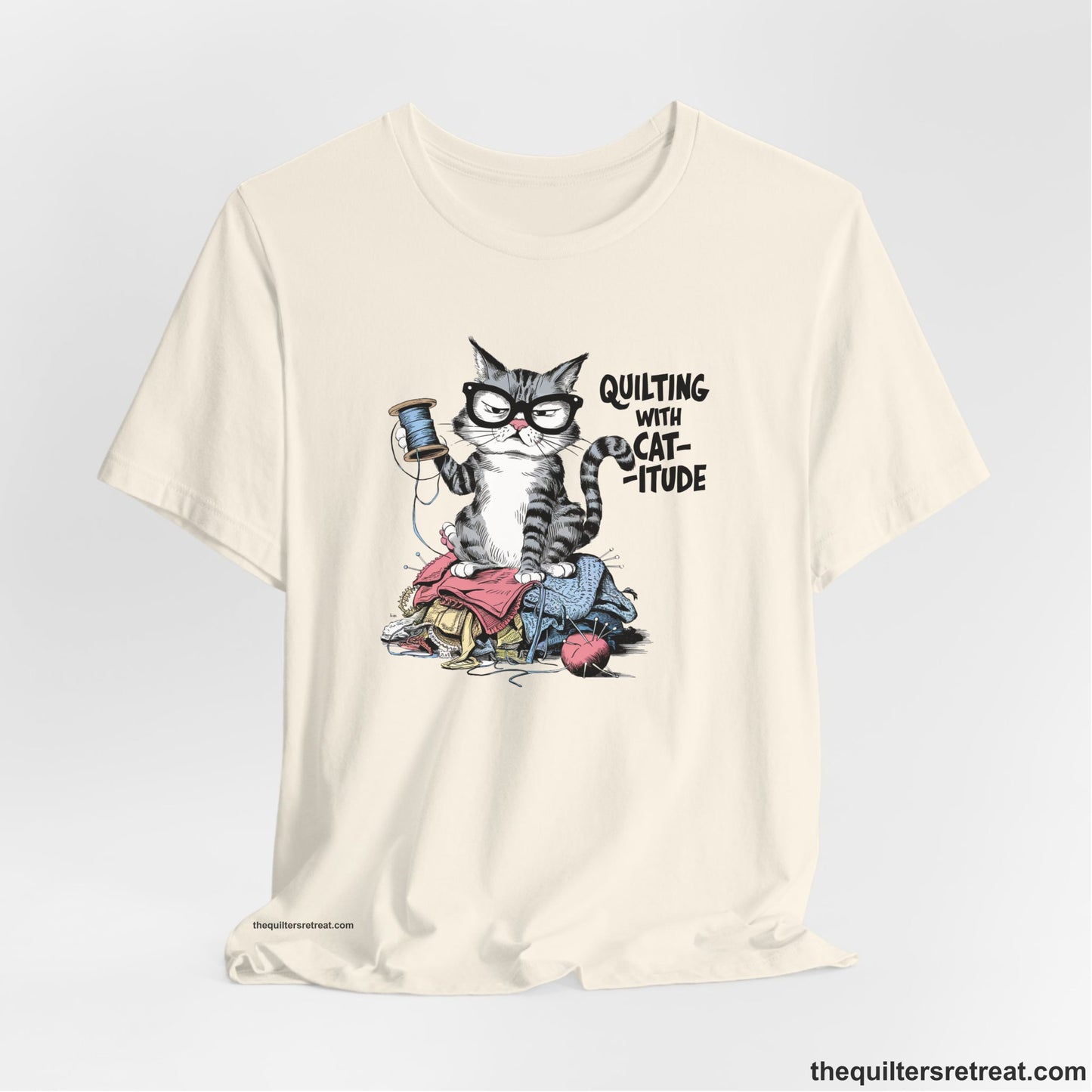 A Natural Short Sleeve T-Shirt with a grumpy cat holding thread spool atop fabric pile with "Quilting with Cat-itude" text