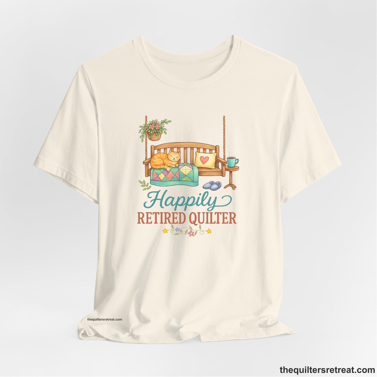 A Natural Short Sleeve T-Shirt with a hanging bench with cozy cat and quilt, surrounded by plants, "Happily Retired Quilter" text