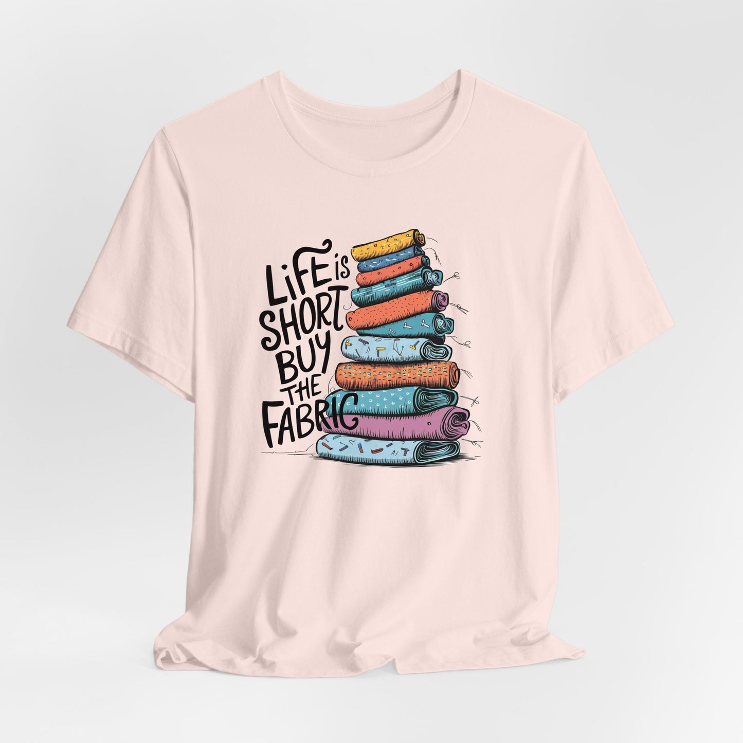 A Soft Pink Short Sleeve T-Shirt with a illustration of colorful fabric rolls with bold "Life is Short Buy the Fabric" text