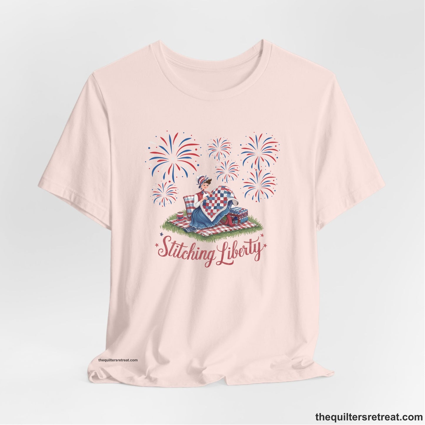 A Soft Pink Short Sleeve T-Shirt with a vintage illustration of woman quilting with fireworks and "Stitching Liberty" text in patriotic style