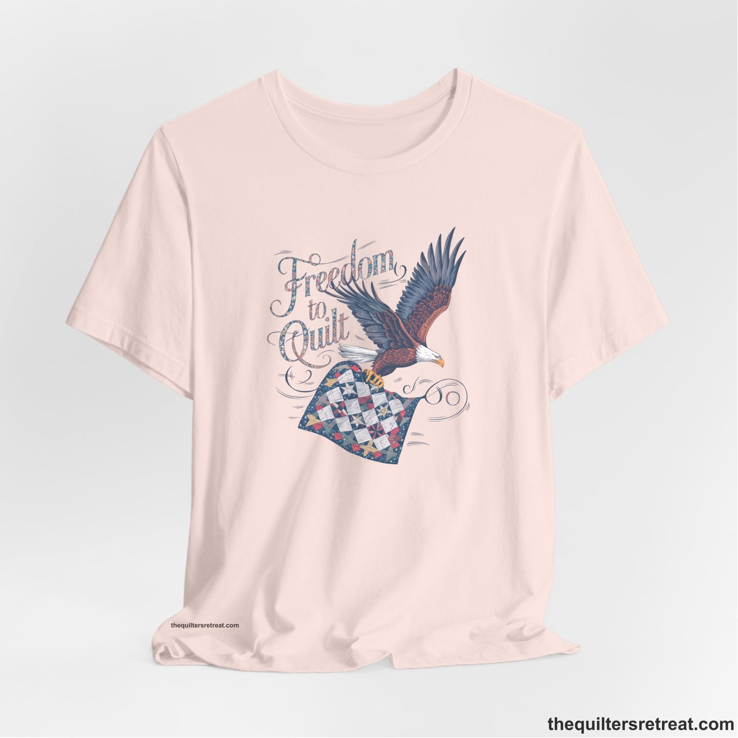 A Soft Pink Short Sleeve T-Shirt with a freedom to quilt text with an eagle carrying a colorful patchwork quilt in vintage style