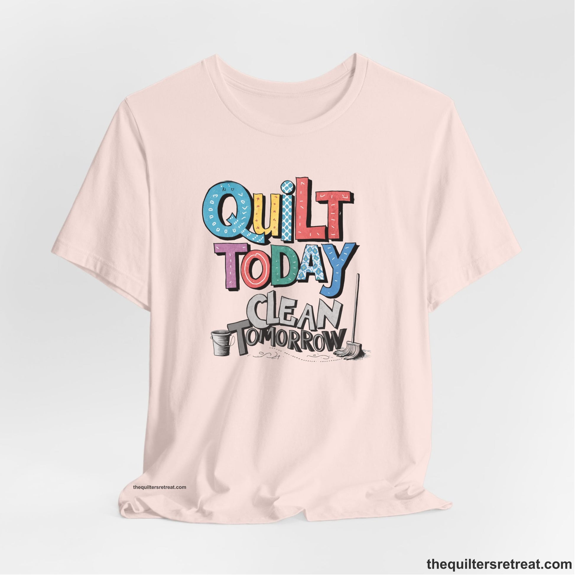 A Soft Pink Short Sleeve T-Shirt with a playful "Quilt Today Clean Tomorrow" text with colorful patterns and cleaning tools illustration
