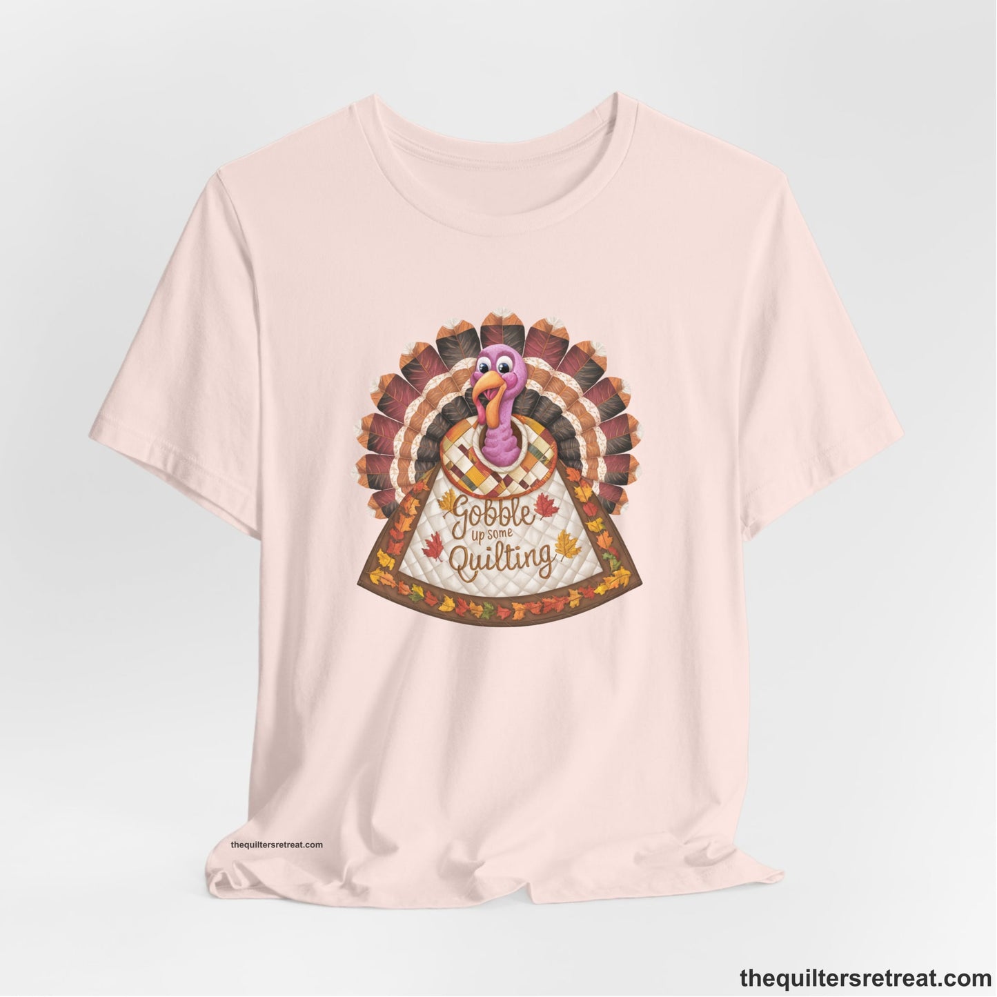 A Soft Pink Short Sleeve T-Shirt with a colorful turkey with quilted feathers and "Gobble Up Some Quilting" text in a playful style