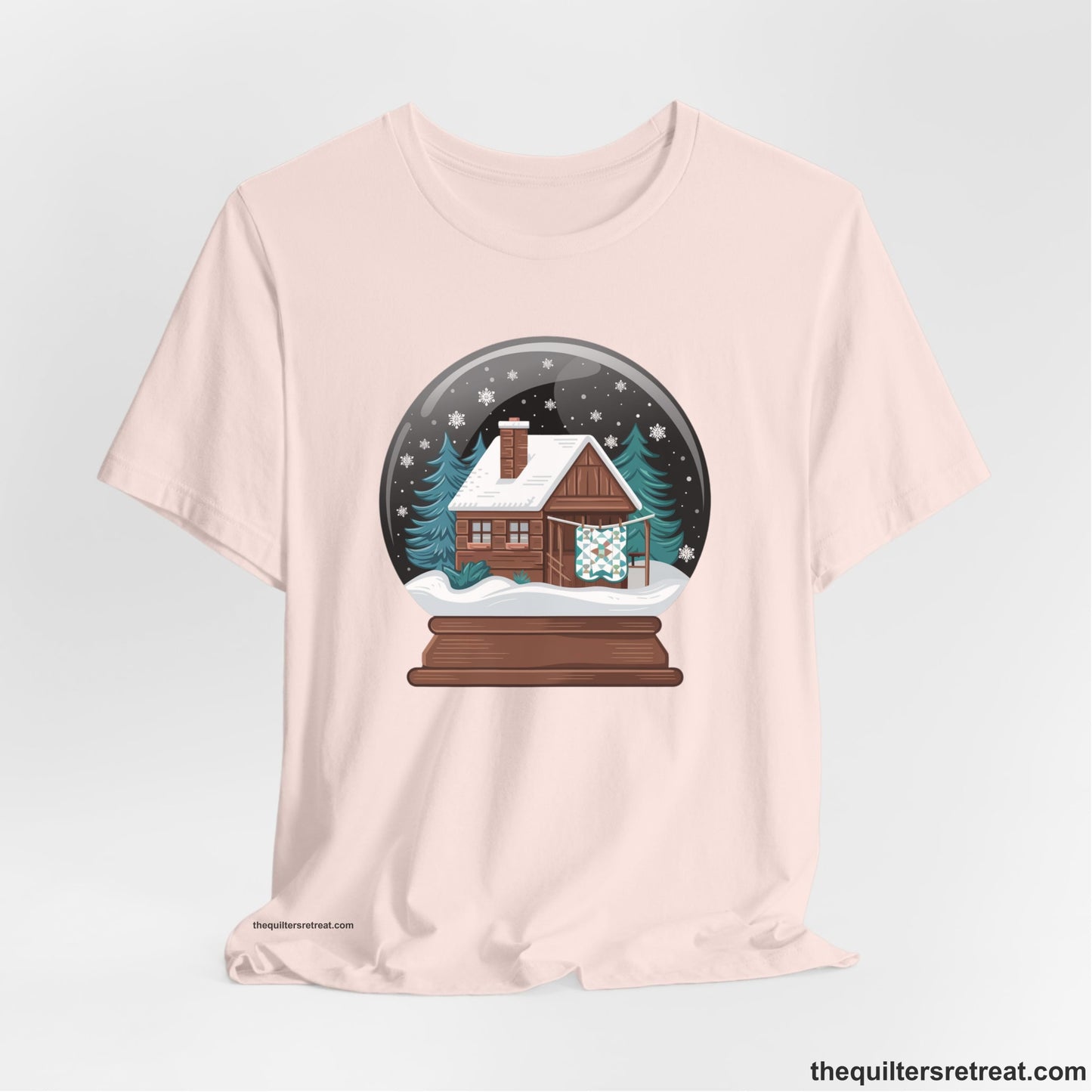 A Soft Pink Short Sleeve T-Shirt with a cabin in a snow globe with snowflakes and trees, in a cozy rustic style