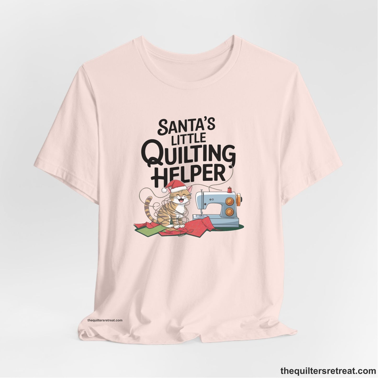 A Soft Pink Short Sleeve T-Shirt with a santa-hat-wearing kitten with a sewing machine and "Santa's Little Quilting Helper" text in playful style