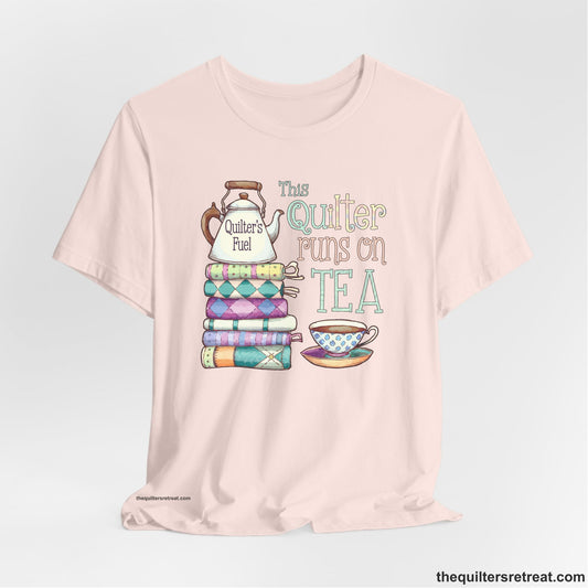 A Soft Pink Short Sleeve T-Shirt with a stack of colorful quilts with teapot and cup, whimsical "This Quilter Runs on Tea" text