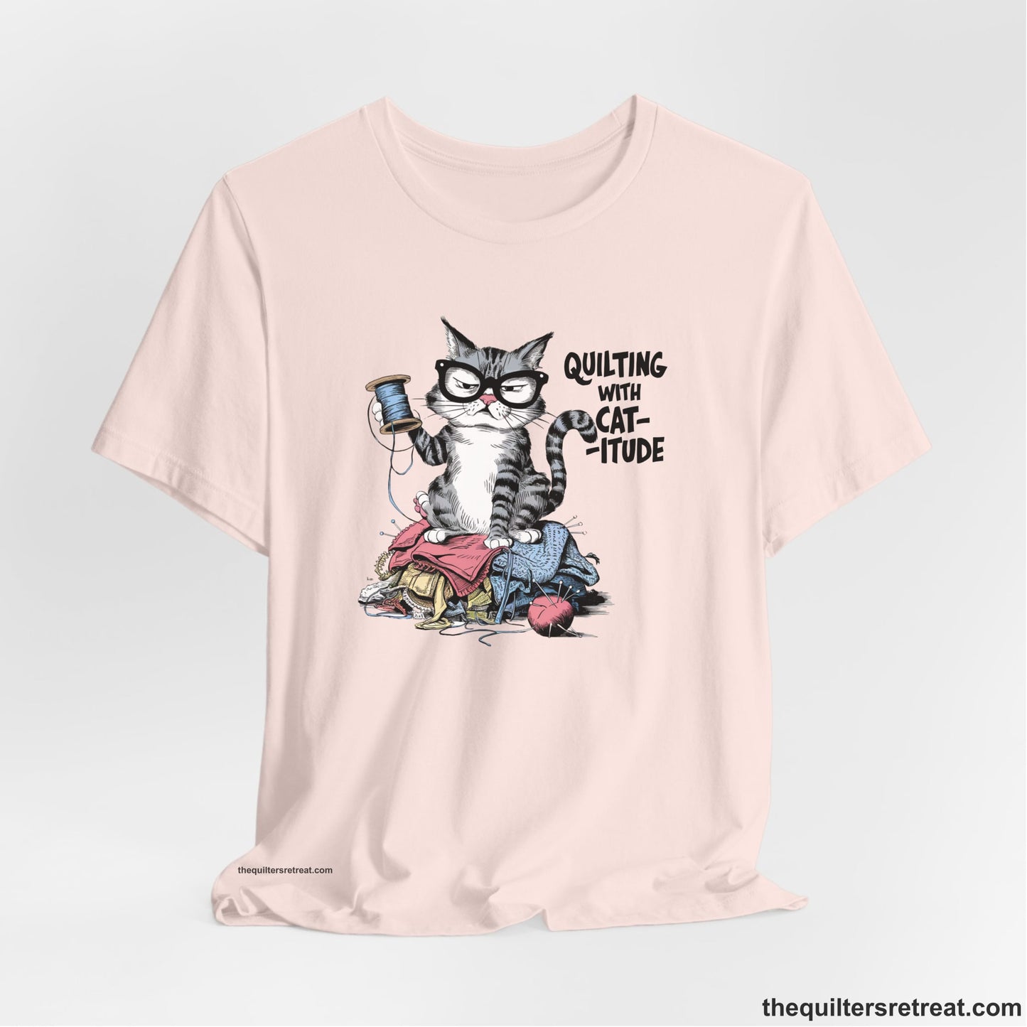 A Soft Pink Short Sleeve T-Shirt with a grumpy cat holding thread spool atop fabric pile with "Quilting with Cat-itude" text