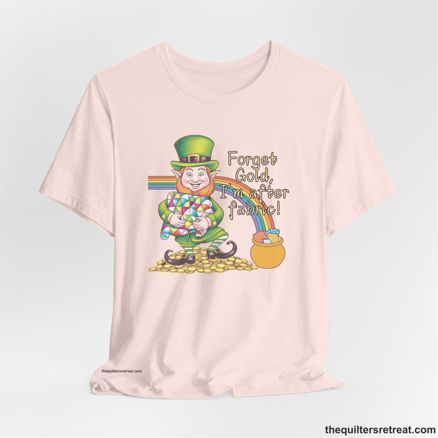 A Soft Pink Short Sleeve T-Shirt with a leprechaun with colorful quilt and rainbow, whimsical "Forget Gold, I'm after fabric!" text