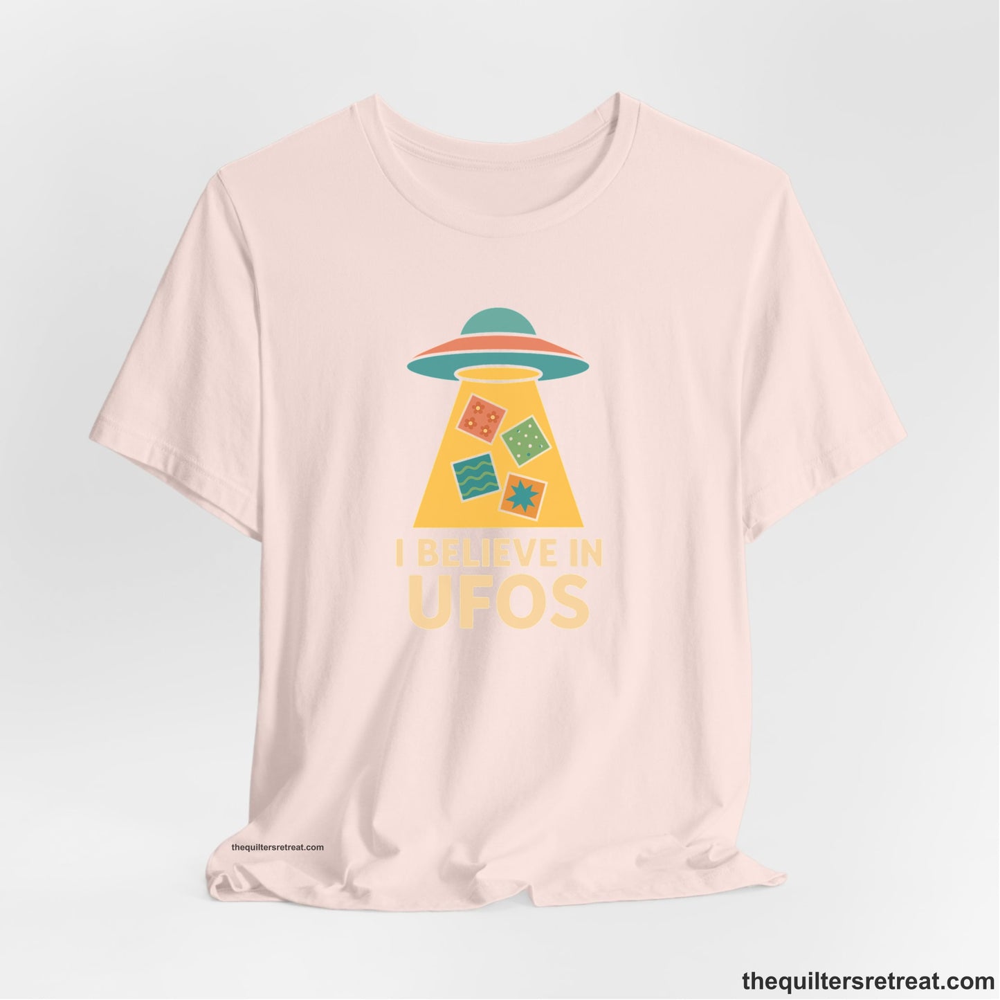 A Soft Pink Short Sleeve T-Shirt with a colorful UFO with quilt patterns and bold "I Believe in UFOs" text in retro style